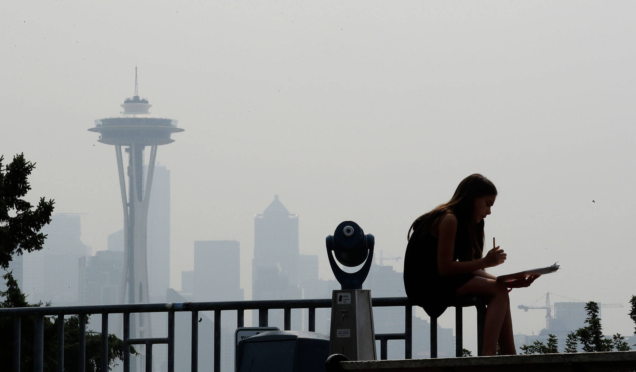 Photo courtesy of The Everett Herald                                A woman works on a drawing next to an unused viewing scope as a smoky haze obscures the Space Needle and downtown Seattle last August as smoke from wildfires moved across the region.